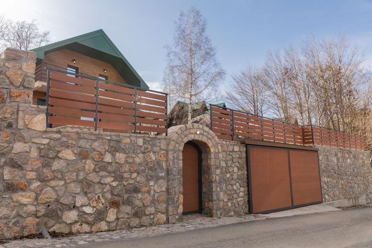 Villa Vučje spolja: drvena fasada sa zelenim krovom, balkon sa drvenom ogradom i visoki kameni zid sa lučnom kapijom uz put.