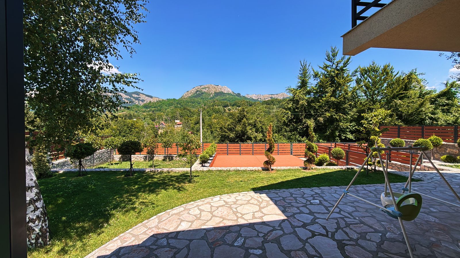 Ljetni pogled iz dvorišta: travnjak, kamenom popločana terasa, crvena drvena ograda i planinski vrhovi pod vedrim nebom.